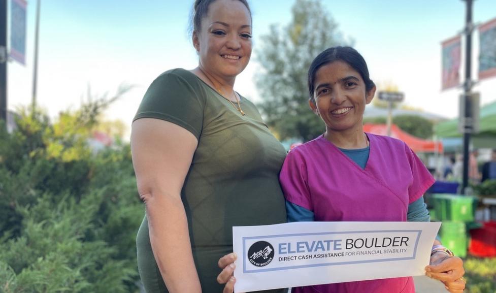 Two people smiling at the camera, the person the right is holding a sign with the Elevate Boulder logo.