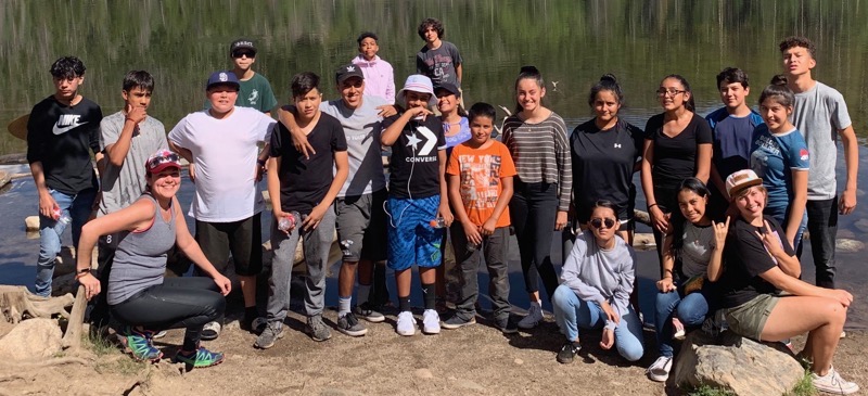 A group of teens and adult mentors pose in front of a lake.