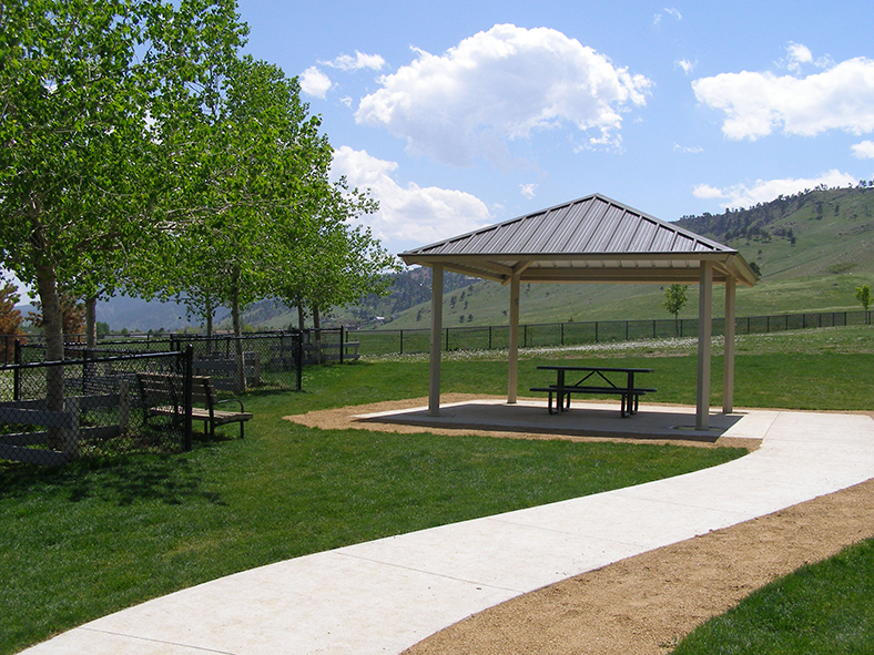 A shade shelter at Foothills Community Dog Park, with a picnic table underneath. It is a sunny day. The shelter is surrounded by green grass and trees, and the foothills are in the background..