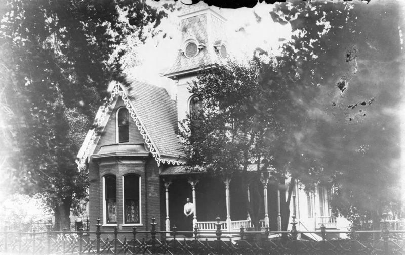 Front view of house with woman standing on porch. Fence in foreground.