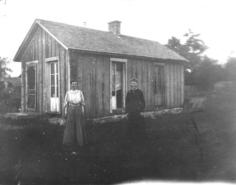 Two women standing in front of a very simple wood frame house