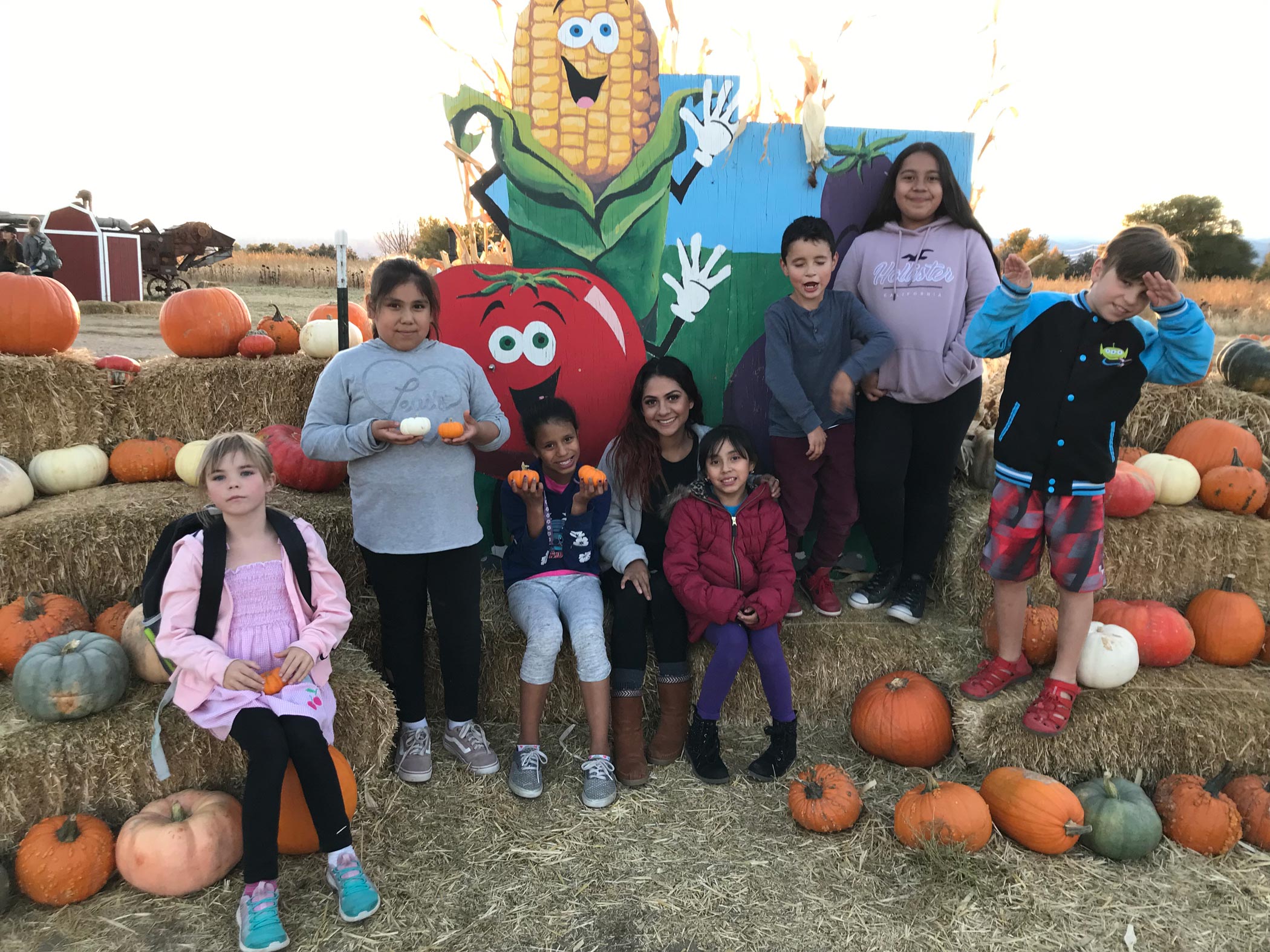 A group of kids pose in front of hay bails and a sign with cartoon vegetables at a pumpkin patch