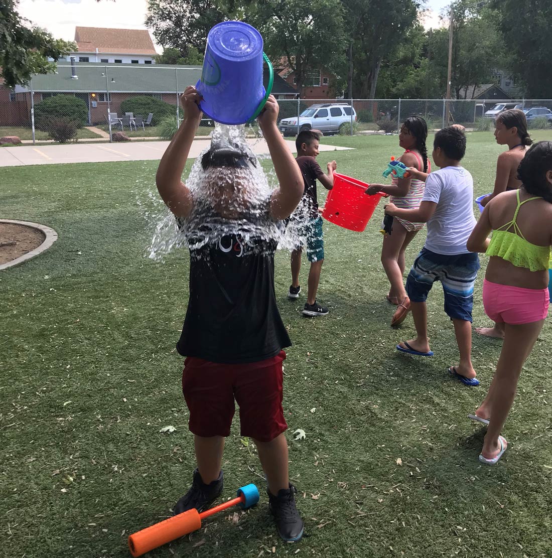 A kid dumps a bucket of water on his head. In the background other kids play with water.