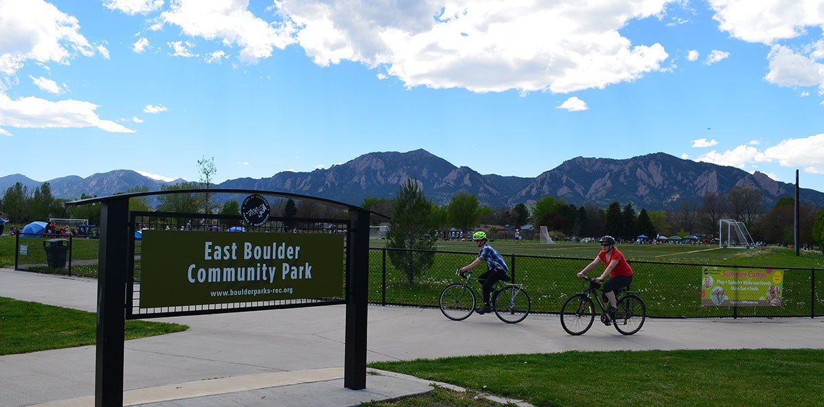 East Boulder Park- two bikers leisurely riding on a maintained path with a soccer field behind them and a view of the flatiron mountains. 