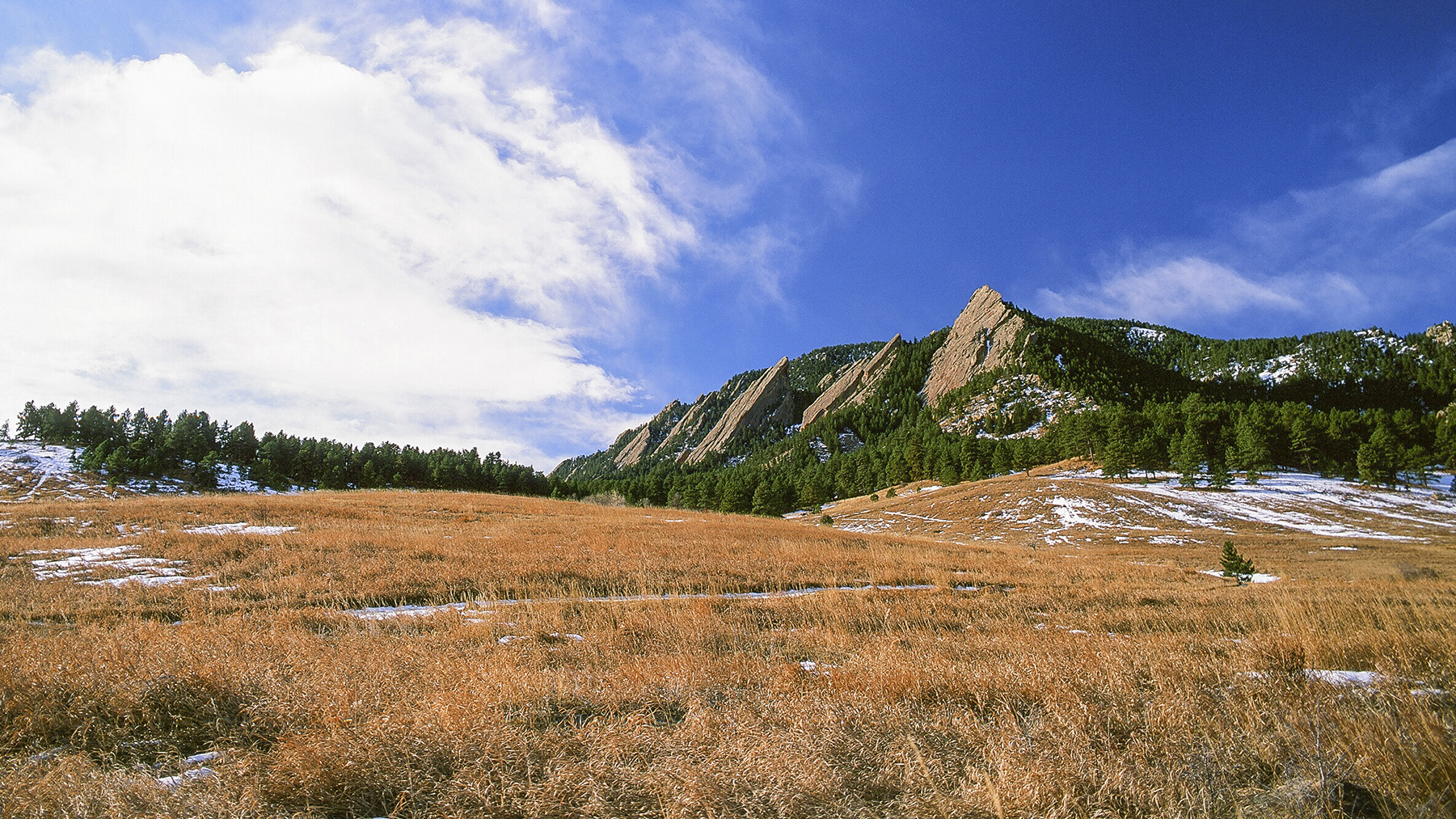 Sun shines on the Boulder Flatirons while clouds cross the sky.
