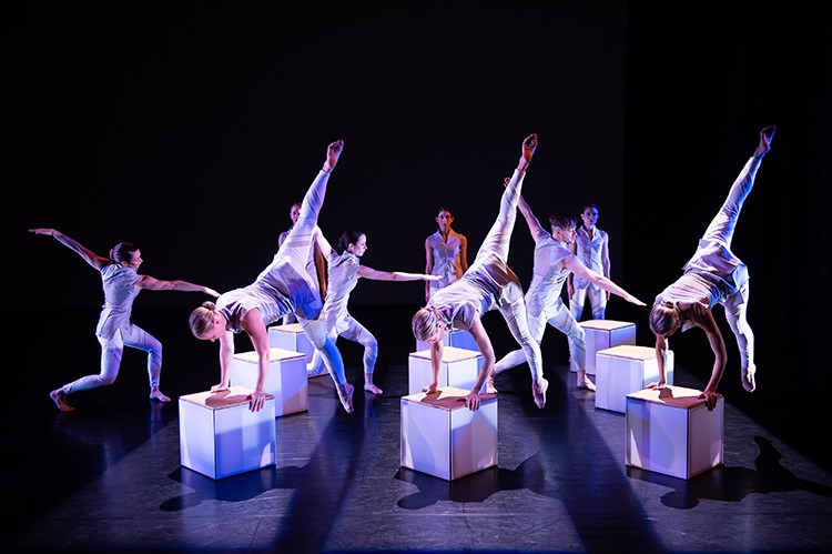 Dance group preforming acrobatic moves on a dark stage, balancing on boxes. 