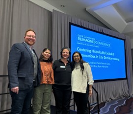 Four people standing in front of a screen on a stage, giving a presentation.