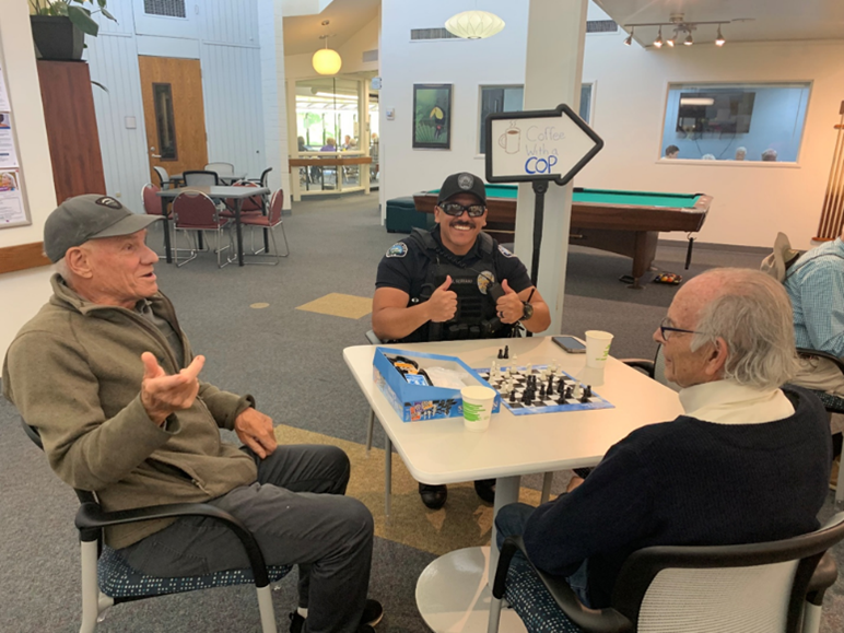 BPD Officer playing a game and having coffee with two older adults at the West Age Well Center. The officer is giving two thumbs up to the camera. 