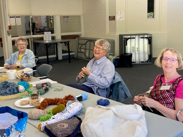 A group of women are sitting at a table covered with different colored yarn and smiling at the camera.