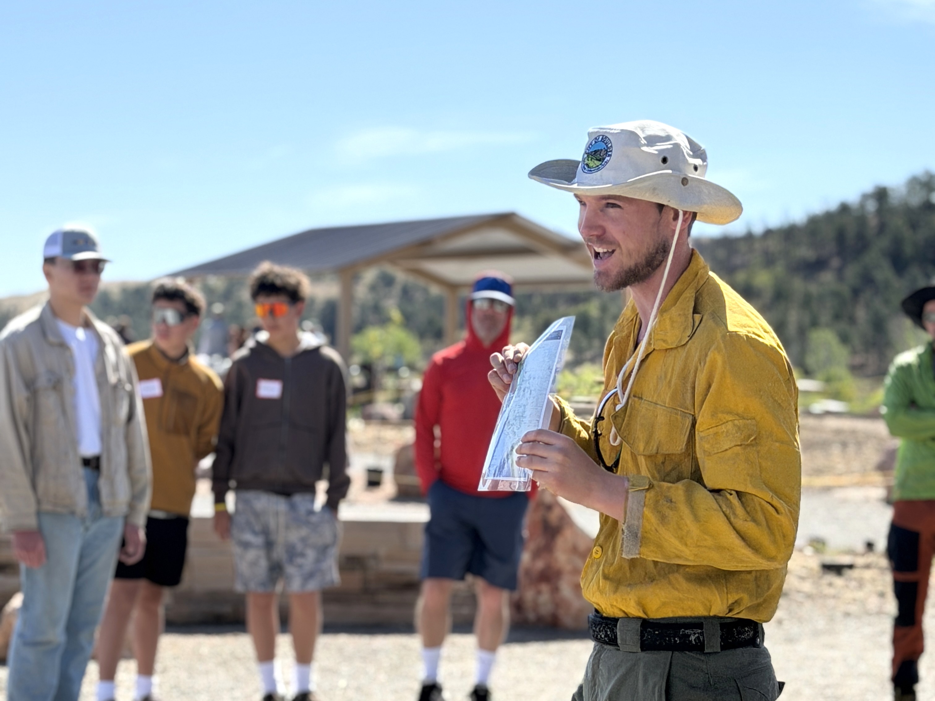 Ranger in wildland fire gear and bucket hat in front of Marshal Mesa holding educational paper and people in background.
