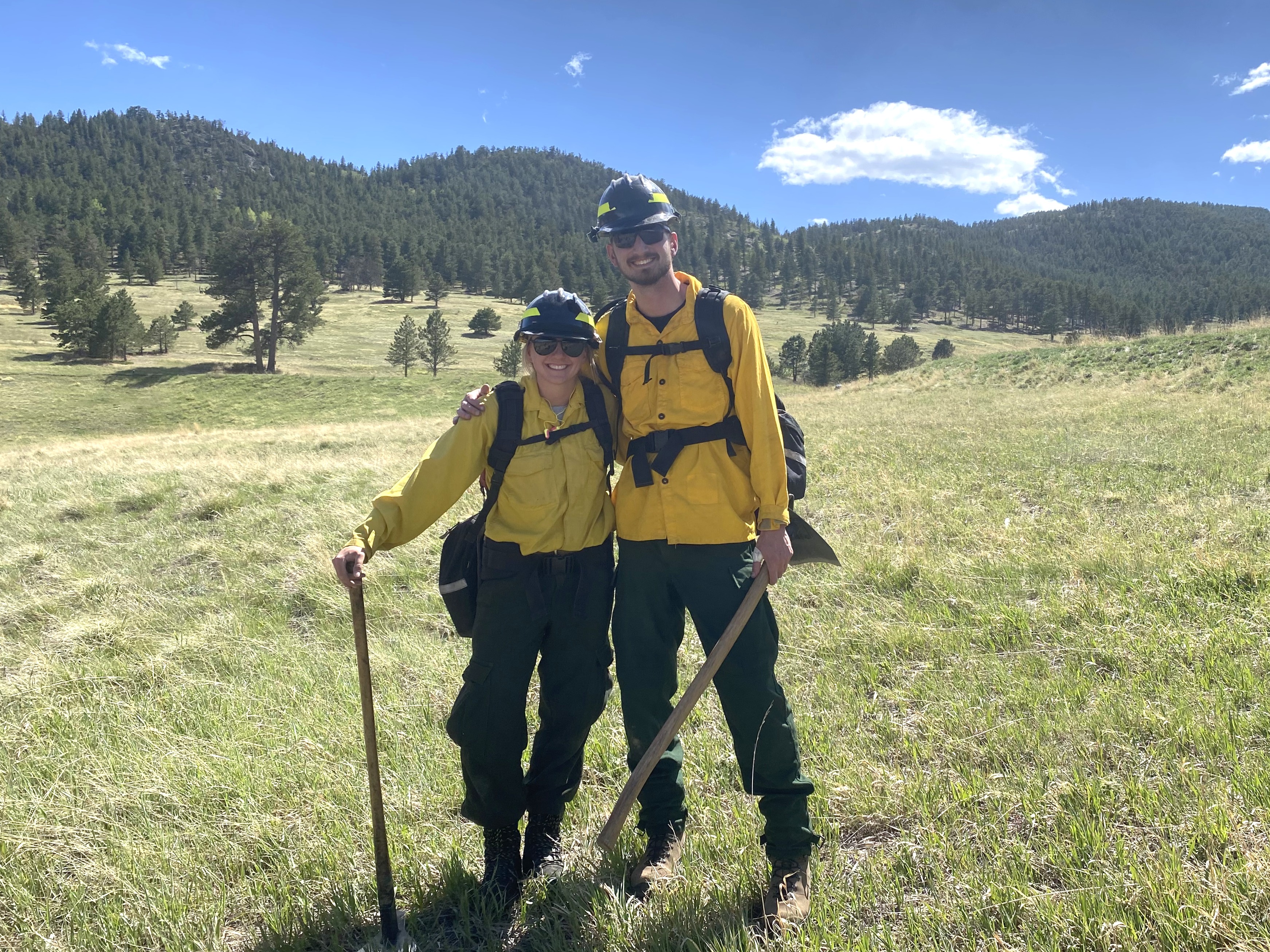 Two rangers in wildland gear with tools in hand and foothills in the background.