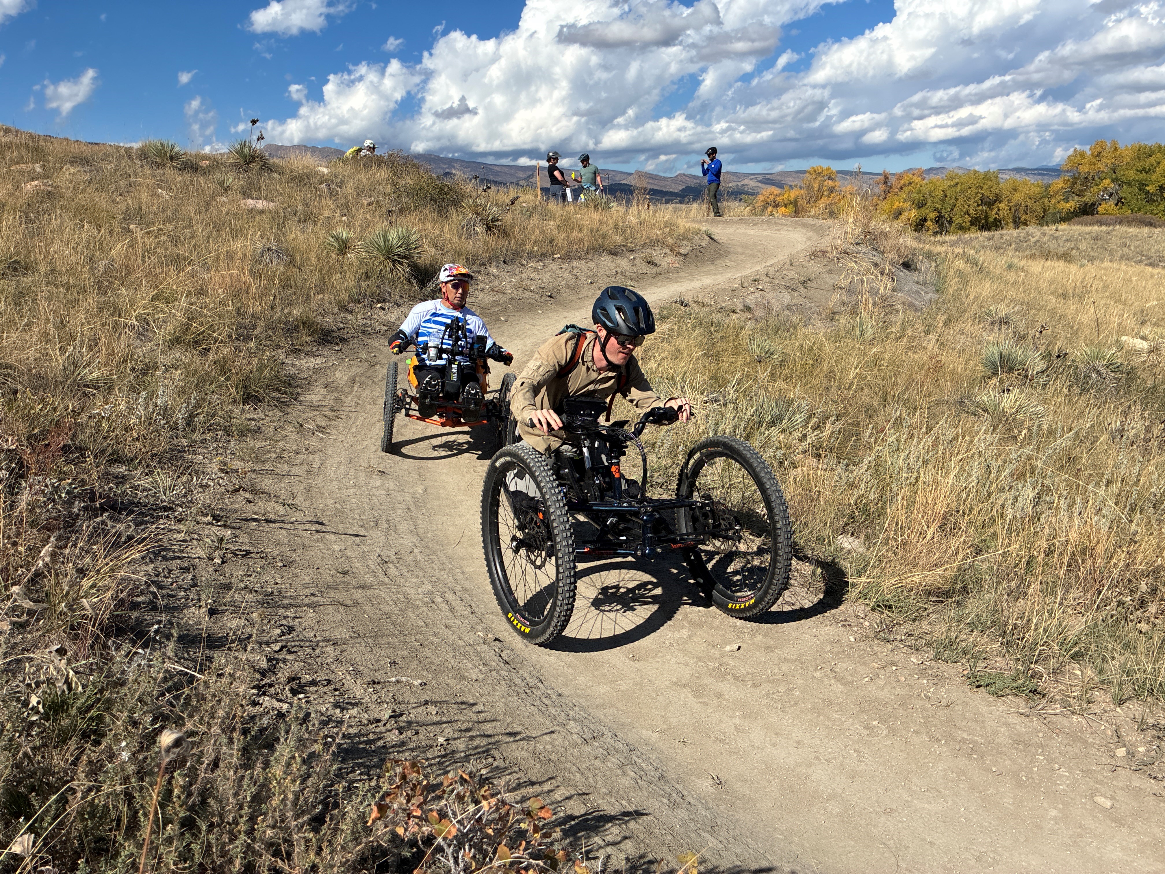 Two people on adaptive mountain bikes riding down a slight hill of a trail.