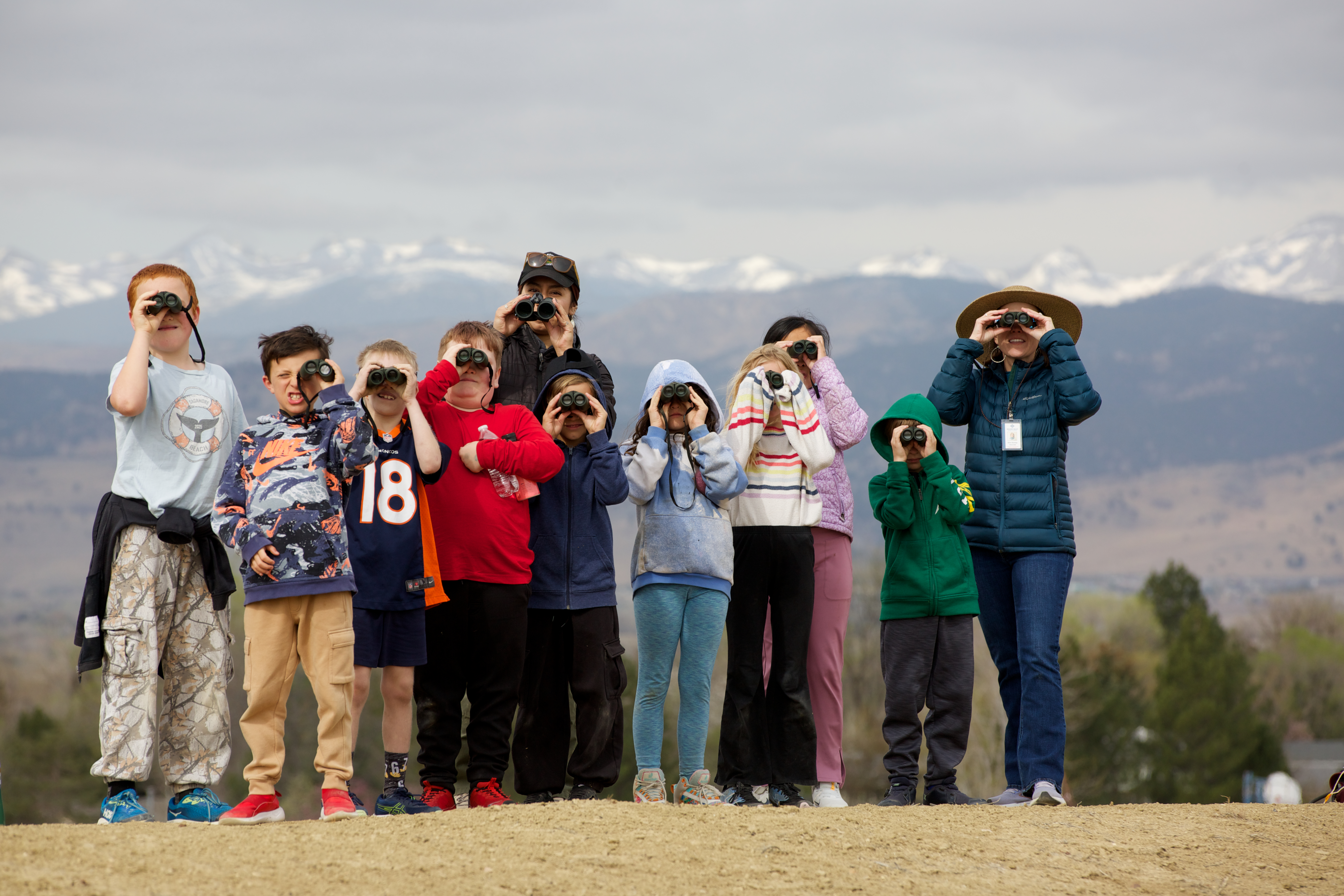A row of young kids standing in front of the mountain range all looking through binoculars.