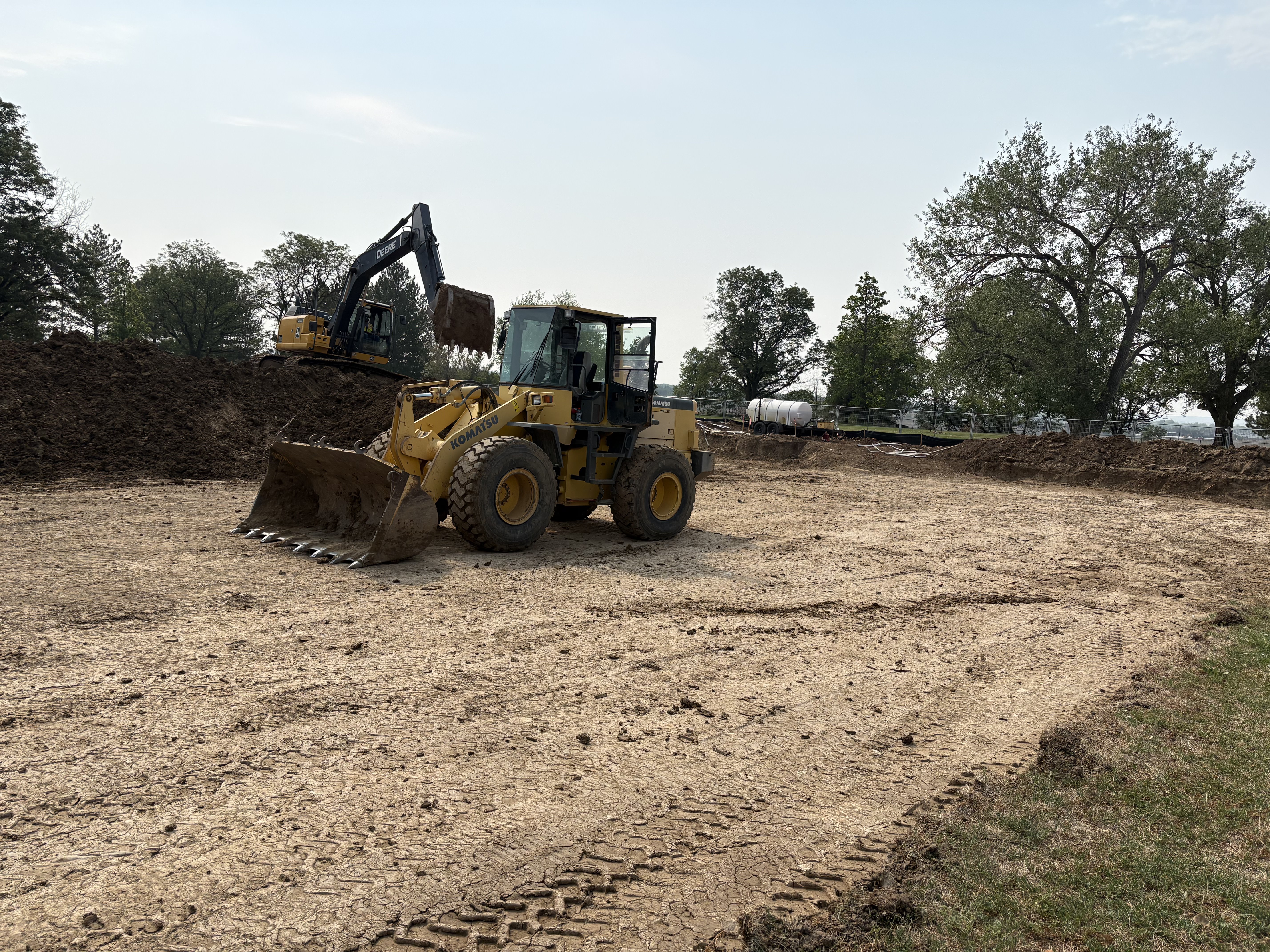 A front-end loader and an excavator move and grade soil at the Tom Watson Park construction site as initial site preparation continues.