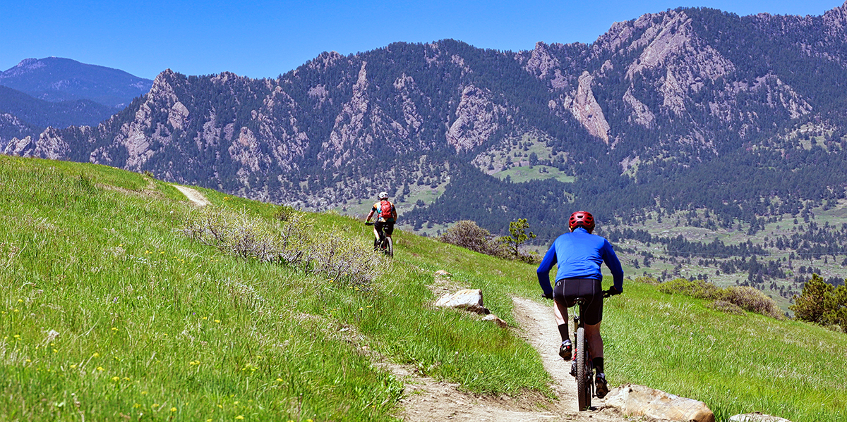 Two bikers on a trail on a grassy mesa overlooking a mountainous landscape