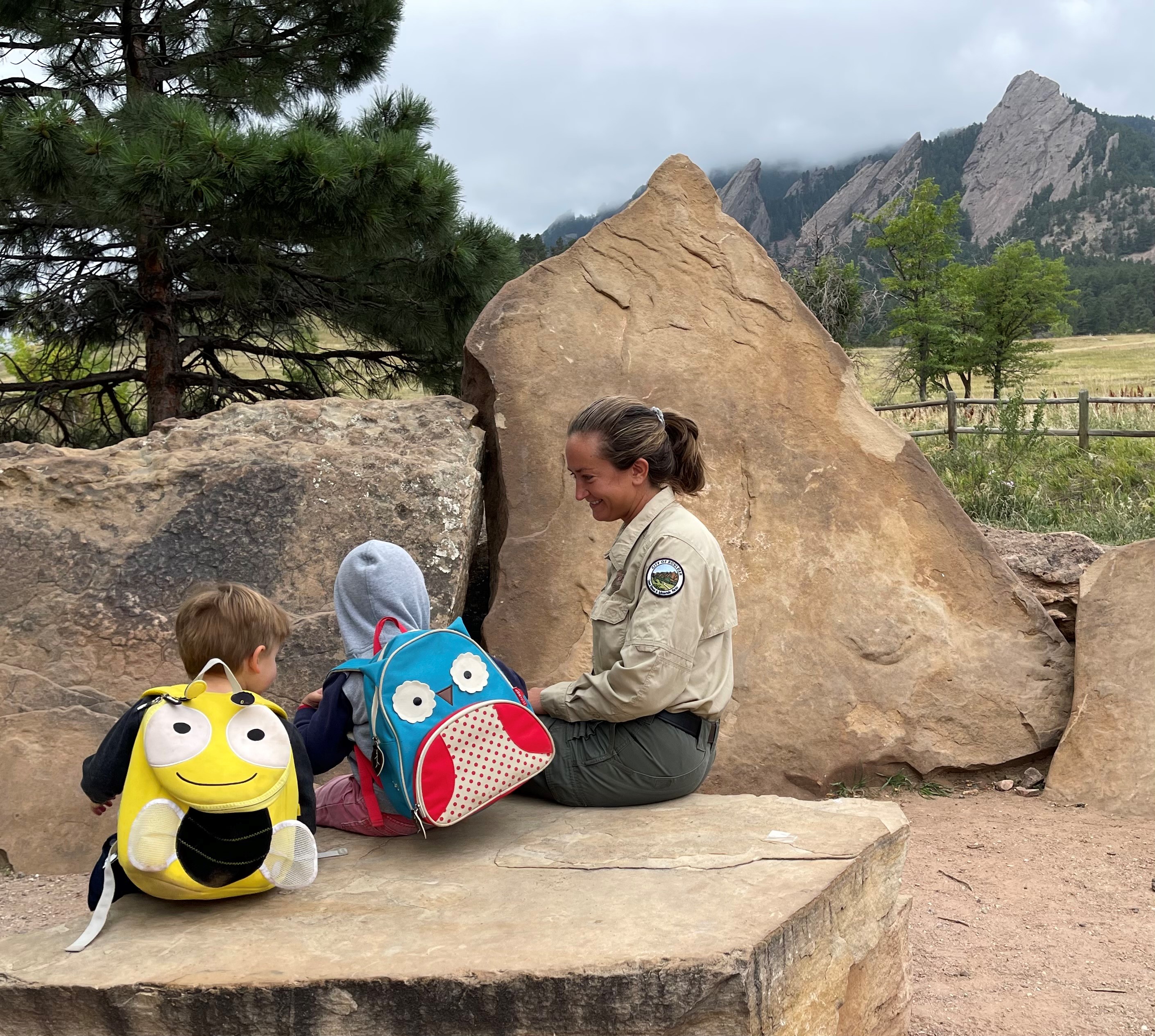 Adult woman in a khaki uniform sits on a rock next to two young kids wearing backpacks like look like animals.