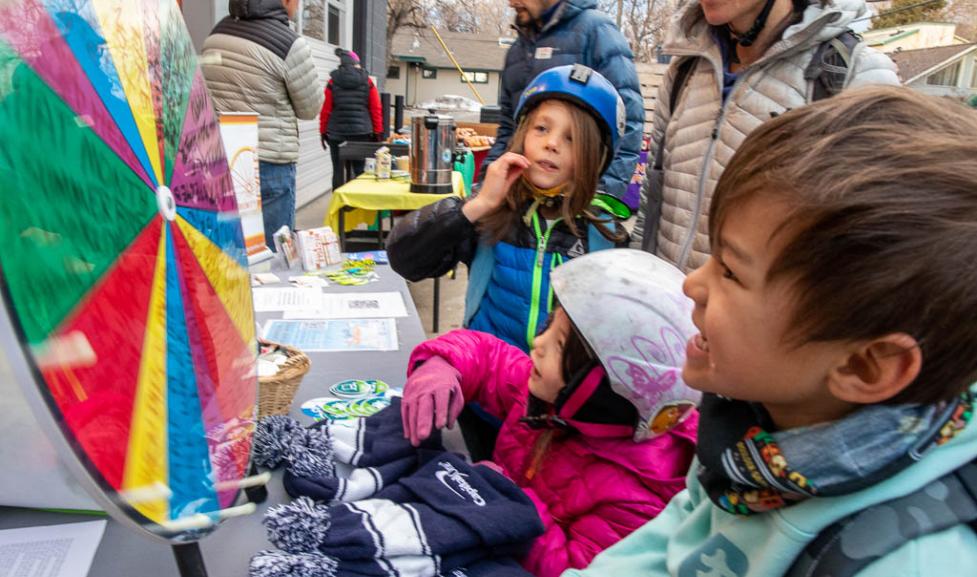 Kids in colorful jackets and helmets outside watch a spinning prize wheel at a table for Winter Bike to Work Day