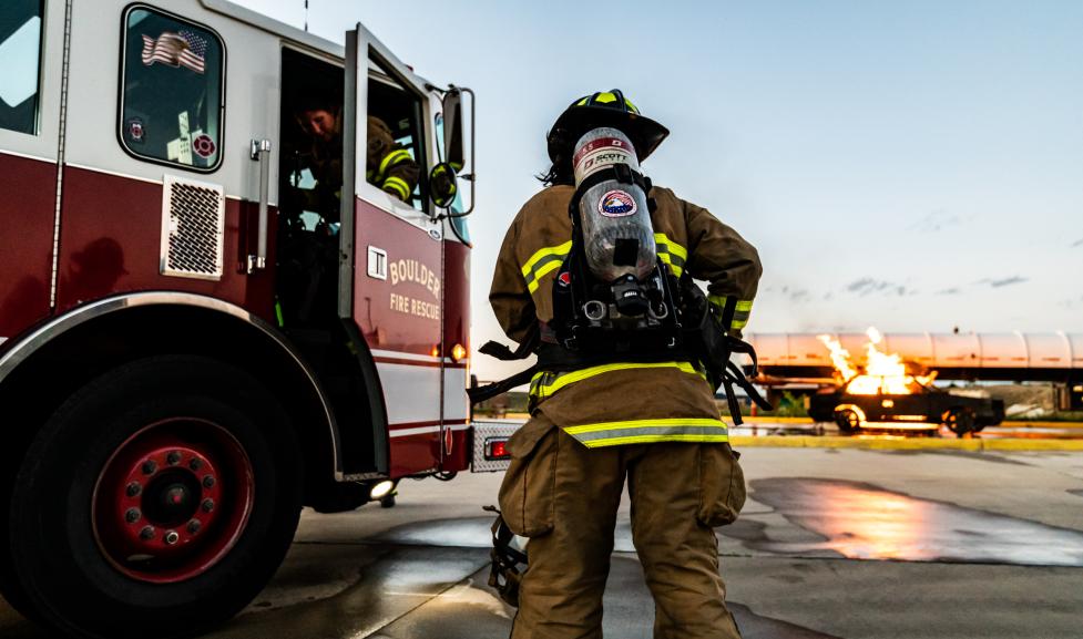 Un bombero en plena concurrencia destaca de pie de espaldas a la cámara. A Boulder El camión de bomberos y rescate está estacionado en el lado izquierdo de la imagen, y el incendio de un automóvil simulado está en el lado derecho de la imagen.