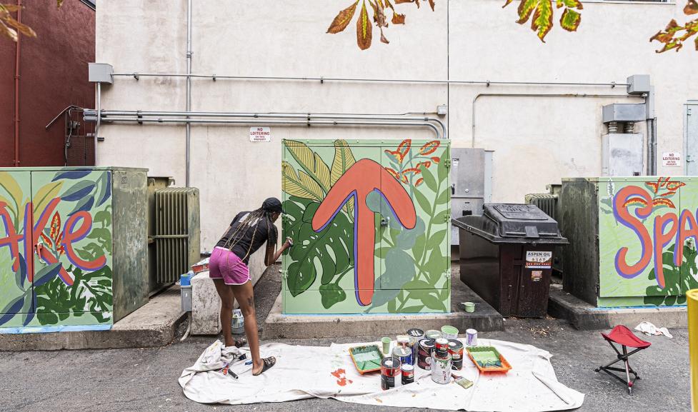 An African American woman with braids tied at the back of her head, wearing a black shirt and pink shorts, bends facing away from the camera to paint a utility box. There are three utility boxes that say 'Take' 'An upwards arrow' and 'Space' or 'take up space'