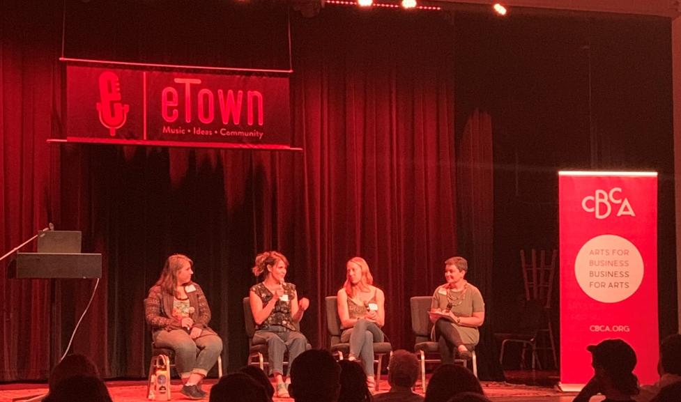 Four women sit panel style on a stage, lit with red tinted stage lights, with a CBCA banner beside them and sign for etown hall above them