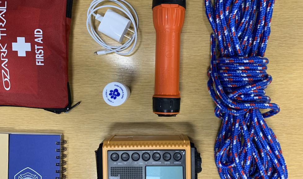 Emergency preparedness tools (First aid kit, charger, flashlight, rope, radio, and notebook) on a table.