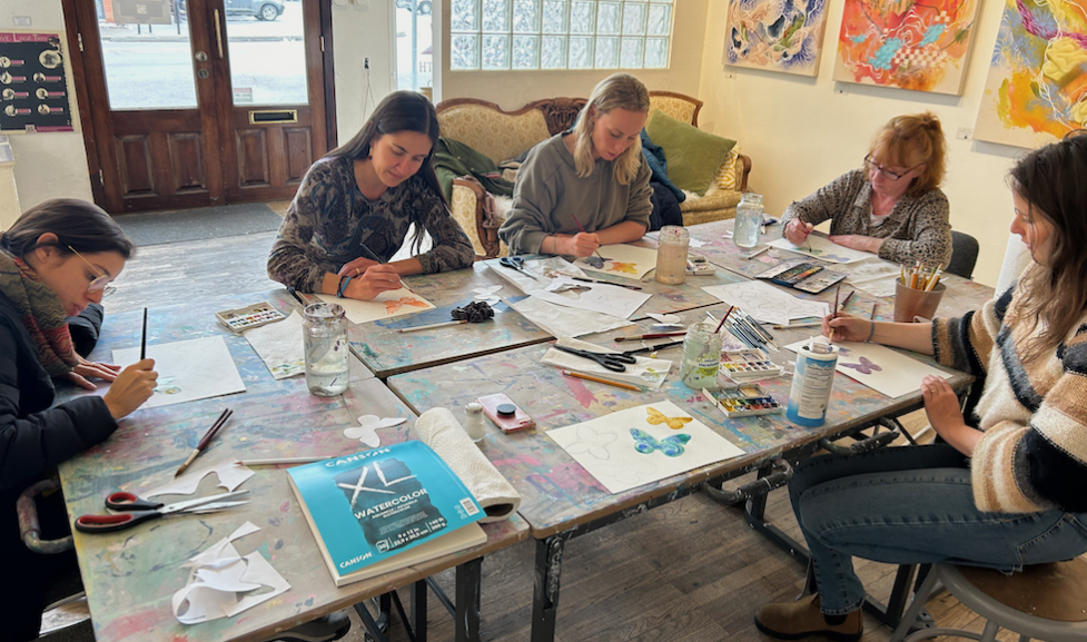 Five women sit around a table painting butterflies in a class for mothers focusing on metamorphosis. The table is old, wooden, and covered in paint spills. The class takes place in a gallery, and there are abstract paintings on the walls.