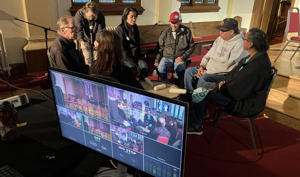 Six people sit in chairs arranged in a circle. A screen in the foreground captures their discussion on film. 