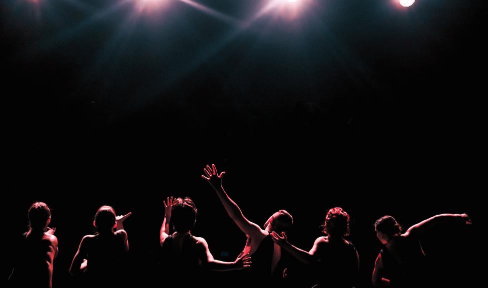 View of the back of stage performers with their arms raised as the lights shine on them. You cannot see the audience because of the stage lights. 