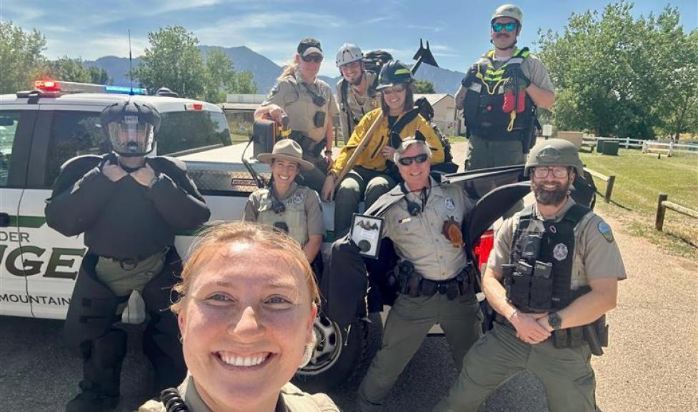 A group of nine rangers posed for a selfie in front of a ranger truck on a sunny day with mountains in the background.  The rangers wear a variety of uniforms representing their diverse roles, including rescue, patrol, education, fire, and medical.