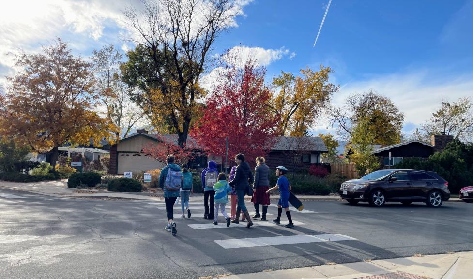 A group of 6 students and 2 parents walking across the crosswalk on Manhattan Drive near Iroquois Drive. A car is stopped at the stop sign, yielding to the group of pedestrians.