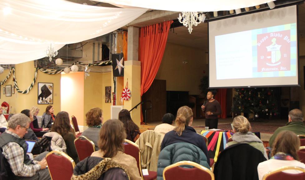 About 30 people in individual red chairs watch a black man present below a projector screen in a festive room.