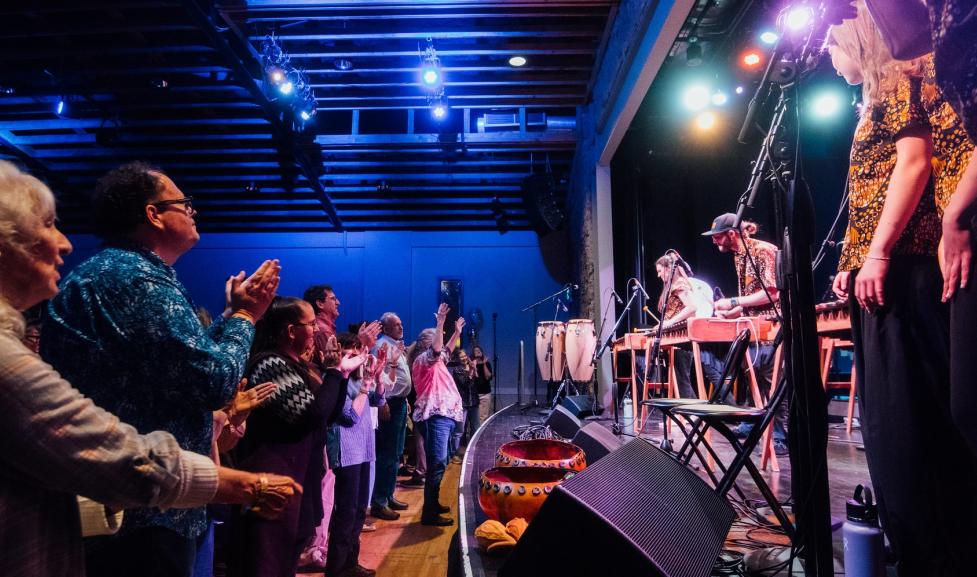 A view from the side of the stage of audience members clapping and enjoy a musical performance with marimbas.