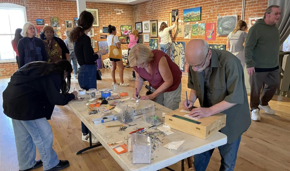 A group of adults do arts and crafts on a table in an art gallery as other adults look at the art on the walls in the background