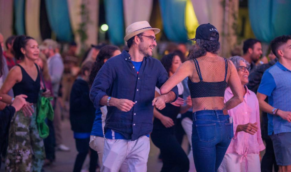 A man and woman samba dance in the forefront as the community dances behind them