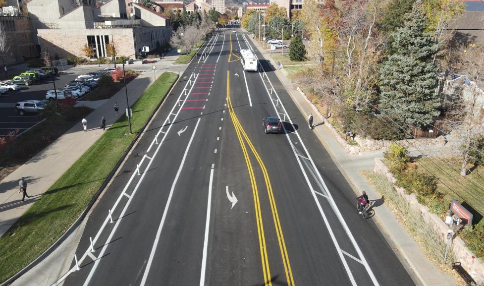 Colorado Avenue shown after 2025 mobility enhancements with wider bike lanes in each direction separated from vehicle lanes by flexible delineator posts, an eastbound bus and right turn lane, an eastbound through lane, a westbound through lane, and turn lanes at the Folsom St and Regent Dr intersections.