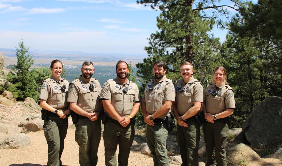 The Urban Ranger Team of six stands on top of Flagstaff Mountain, framed by trees and blue skies, with a view of Boulder in the background. 