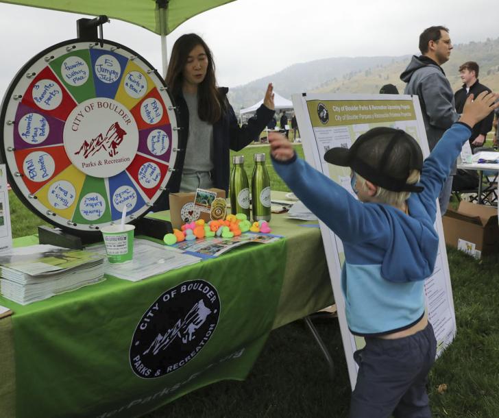 Parks and Recreation table with colorful spinning wheel and a young boy in front of the table with his hands in the air excited about winning something. 