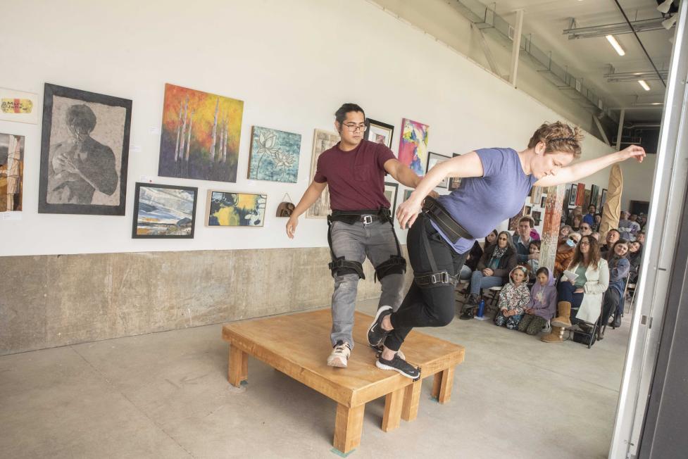 Live art performance of an individual balancing another person off a wooden ledge.