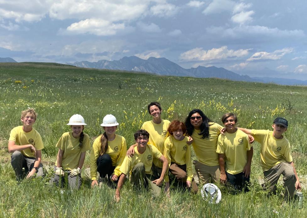Group of teenagers on a trail in a meadow filled with flowers posing for a picture with the mountains in the background.