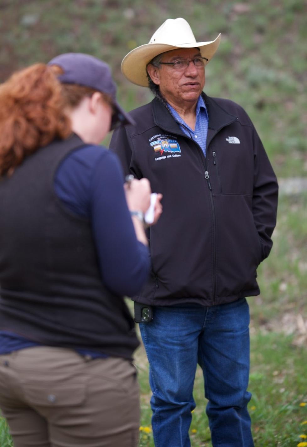 Fred Mosqueda is interviewed by Jessica Yaquinto, who writes in a notebook with her back to the camera. Mosqueda stands beyond her, speaking while looking into the distance, with an out‑of‑focus green field behind them.