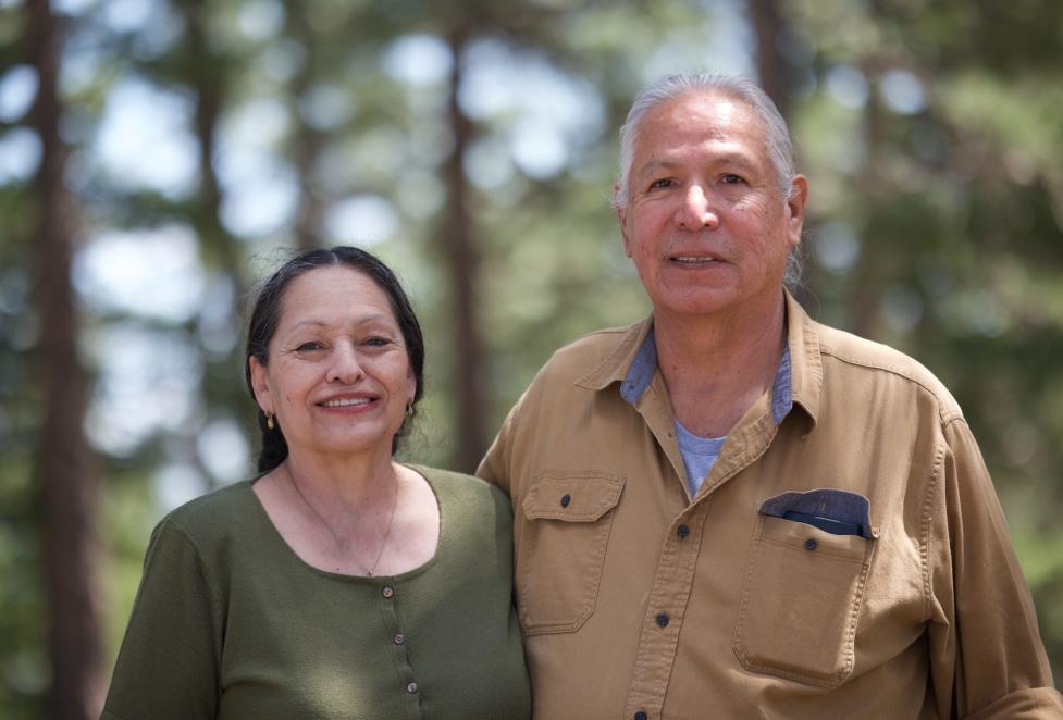 Portrait of married couple Frank and Pauline Medicine Water standing side by side with arms around each other, smiling before an out‑of‑focus forested background.
