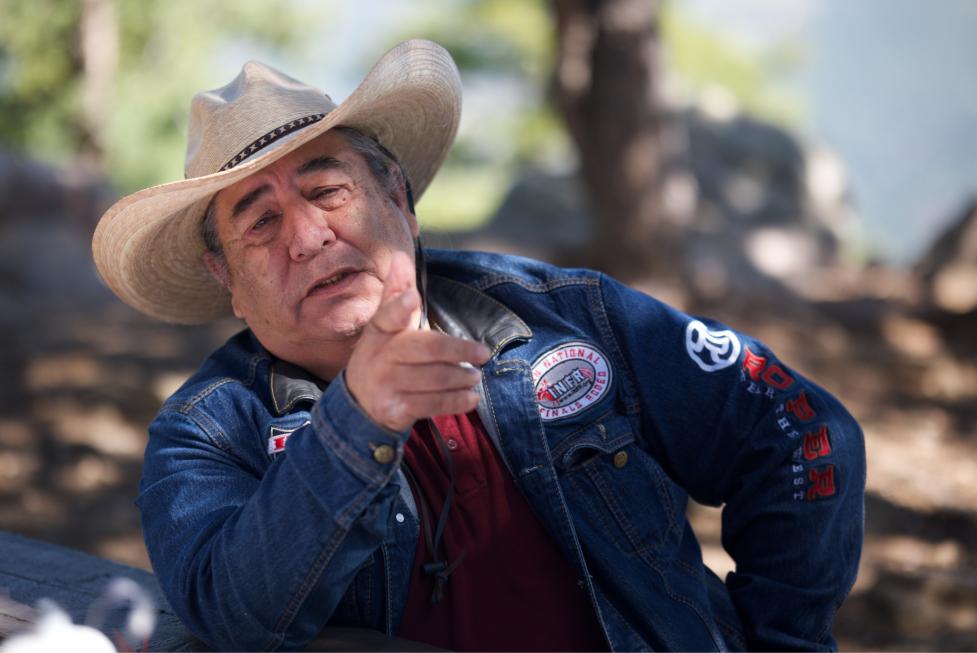 Candid portrait of Chester Whiteman seated at a wooden picnic table, speaking and pointing behind the photographer, with a wooded background out of focus.
