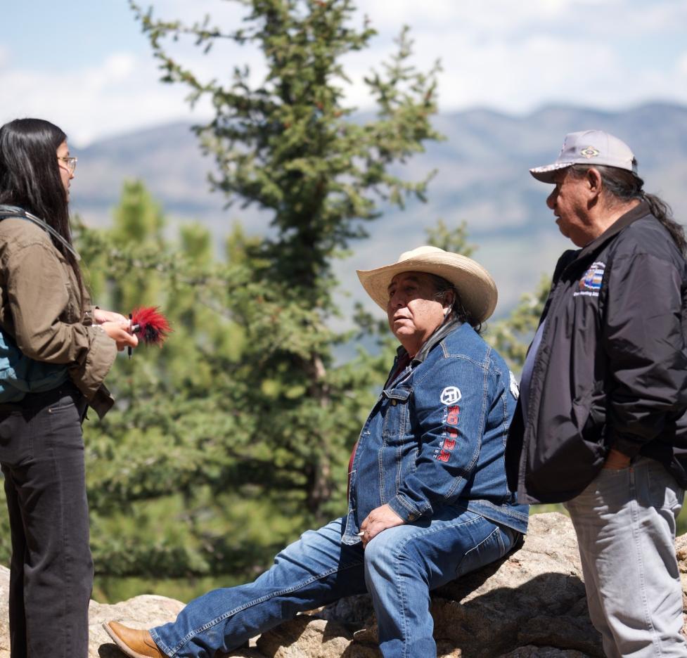 Side view of Mr. Chester Whiteman and Mr. Roy Dean Bullcoming being interviewed by Joanie Finch, who holds a recorder while speaking with Mr. Whiteman seated on a boulder.