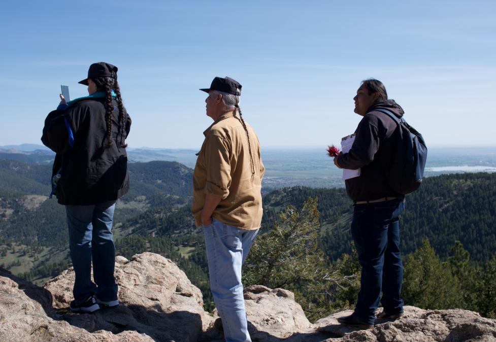 Mr. Frank and Mrs. Pauline Medicine Water stand with ethnographer Reshawn Edison on large rocks overlooking distant mountains.
