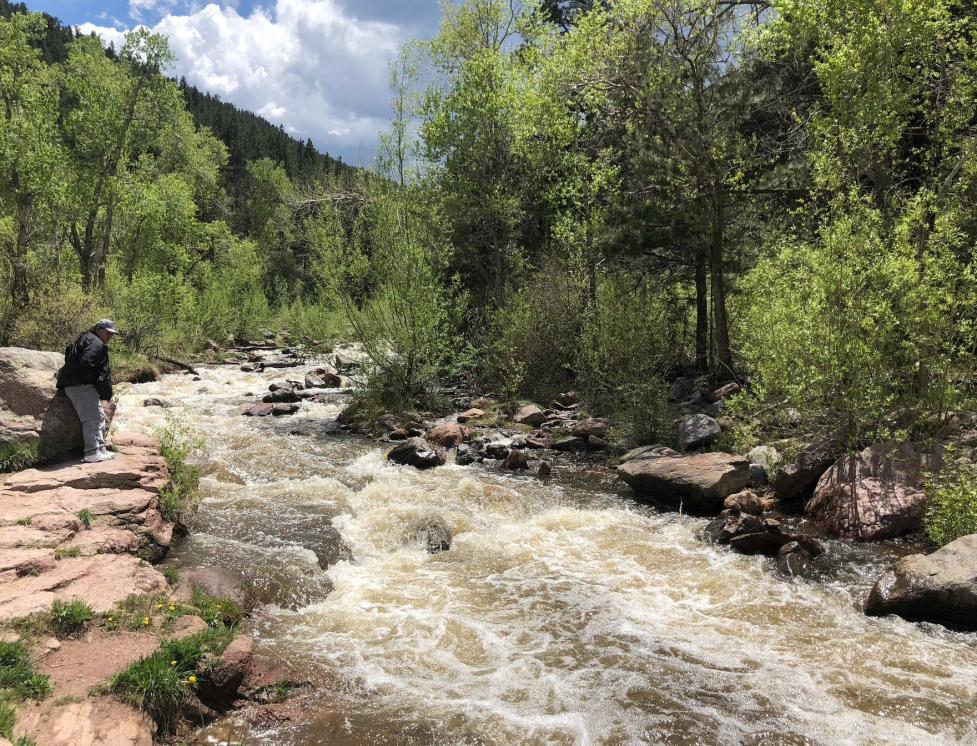 Mr. Roy Dean Bullcoming stands on a rocky outcrop beside a fast‑moving stream, with forested banks and a blue sky with large white clouds.