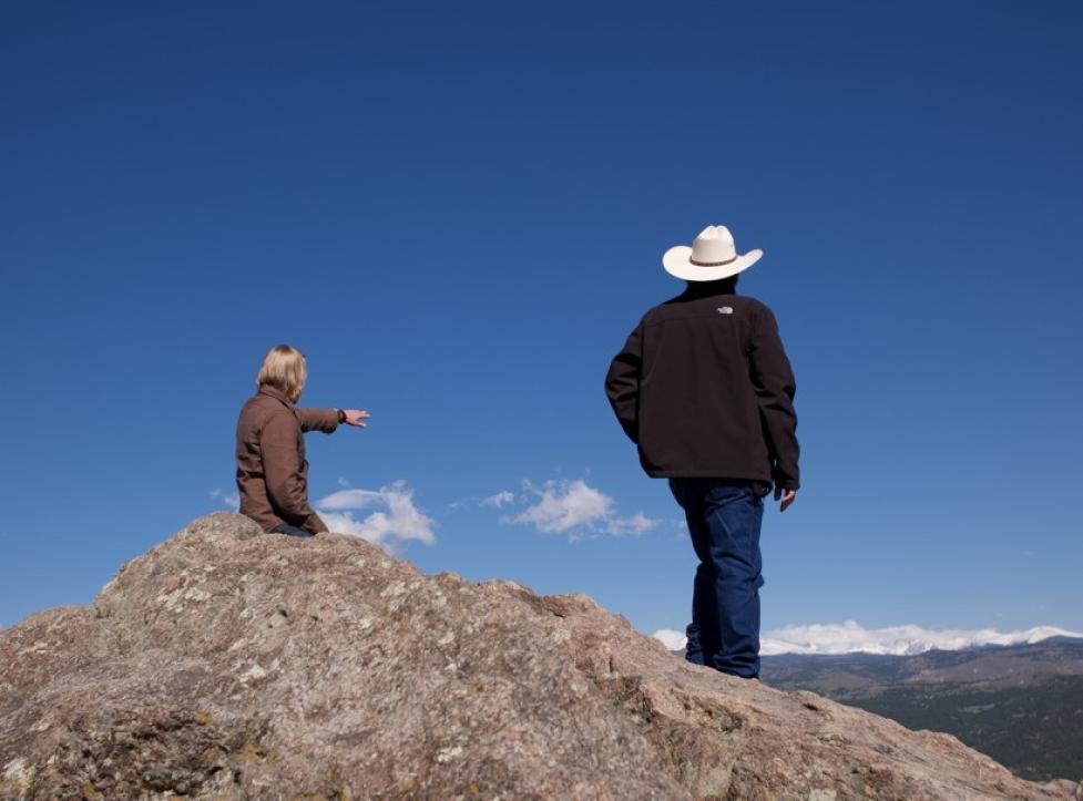 David Ford and Fred Mosqueda stand with their backs to the camera at a Boulder OSMP overlook, partly hidden by a large boulder, as David points toward distant green and snow‑capped mountains beneath a clear blue sky.