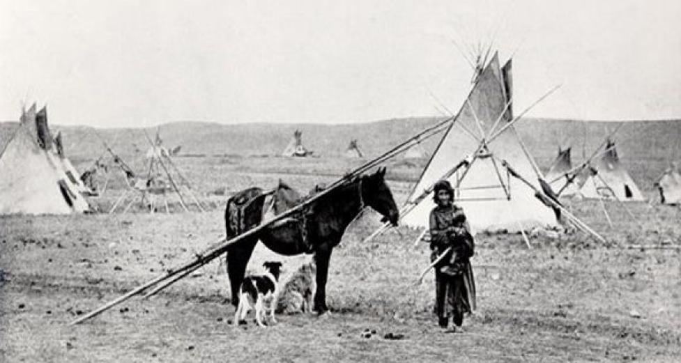 Black‑and‑white image of a Comanche camp with tipis, a person in the foreground, and a horse with a travois and a dog beside it.