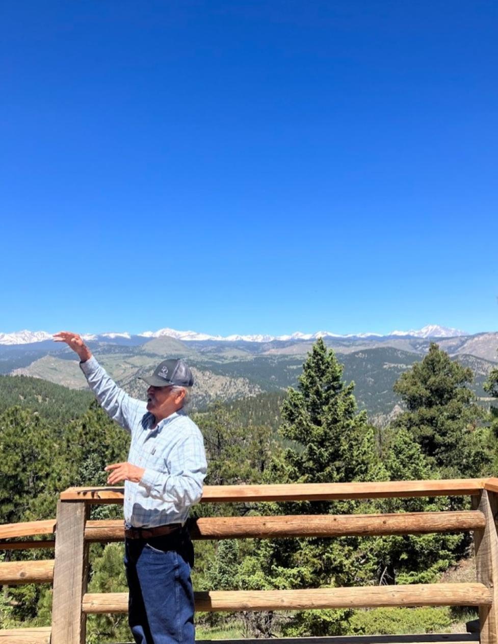 Mr. Leonard Chibity stands at an overlook in Boulder OSMP, shown in profile speaking to someone off‑camera, with conifer‑covered mountains under a clear blue sky behind him.