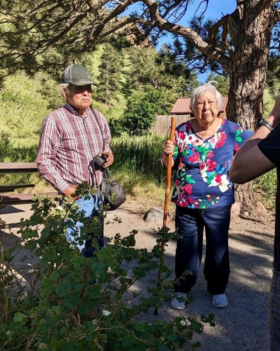 Leonard and Rosemary Chibity stand facing forward in a wooded area, listening to someone off‑camera.