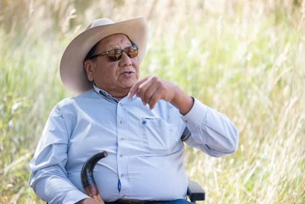 Portrait of Mr. Jhon Goes in Center speaking to someone off‑camera, seated with tall grasses blurred behind.